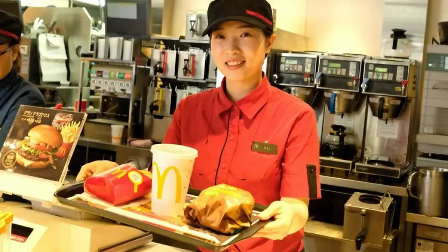 A McDonald's employee in a red uniform and cap smiles while handing a tray of food with a burger, fries, and a drink to a customer at the counter. The kitchen and menu displays are visible in the background.
