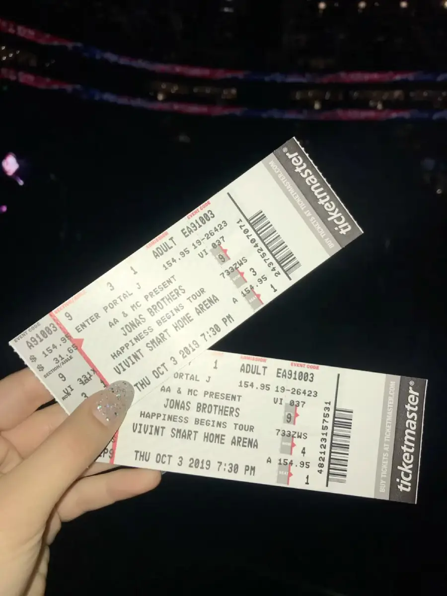 A hand with glittery nails holds two Jonas Brothers concert tickets for the Happiness Begins Tour at Vivint Smart Home Arena, dated October 3, 2019, with a dark, blurred stadium in the background.