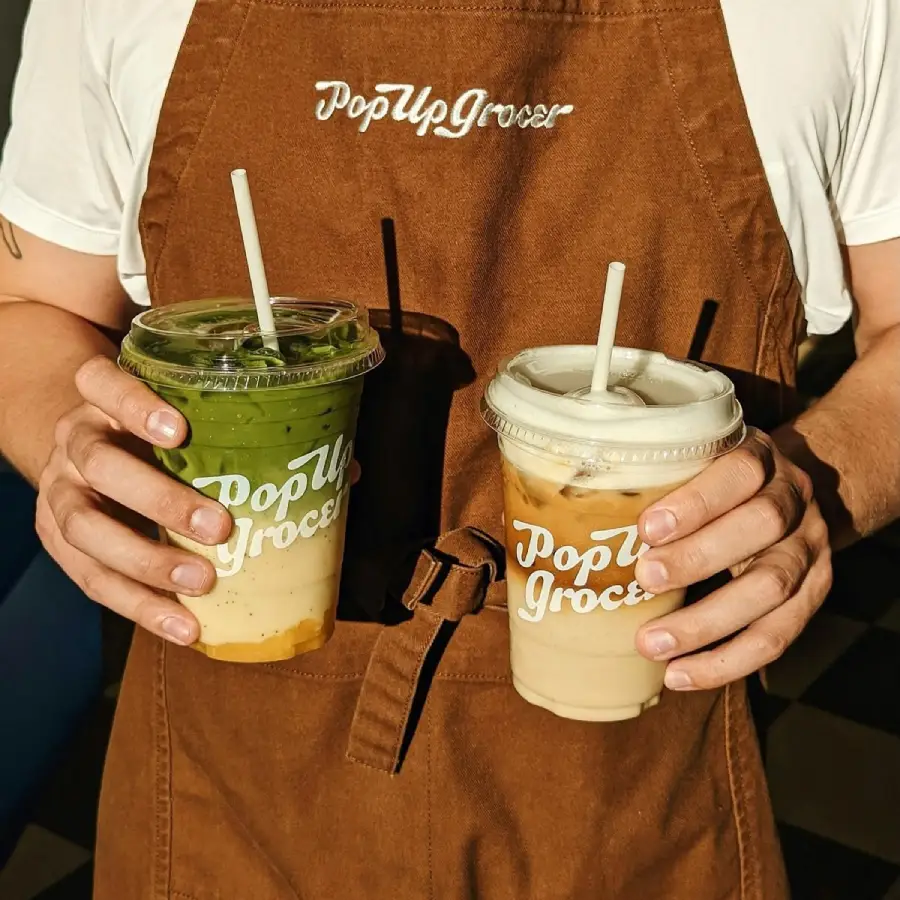 A person wearing a brown "Pop Up Grocer" apron holds two drinks in plastic cups with straws: one green and one beige, both in sunlight.
