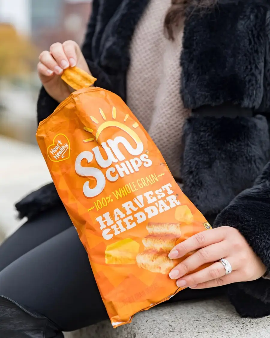 A person wearing a black coat and beige sweater holds an open bag of SunChips Harvest Cheddar, taking a chip out with one hand. The bag is orange and features the SunChips logo and cheddar imagery.