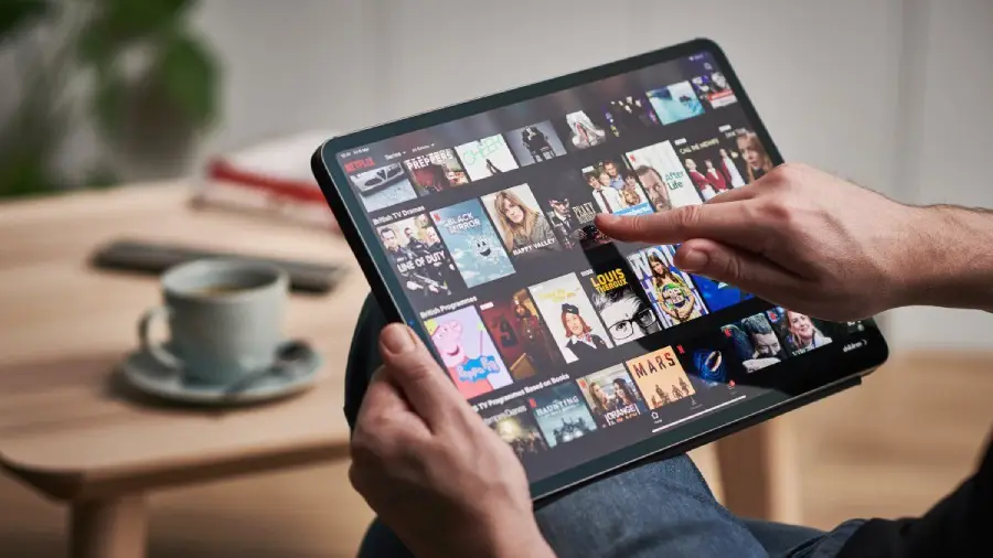 A person holds a tablet and browses through a streaming service interface, scrolling through various TV shows and movies. A cup of coffee sits on a table in the background.