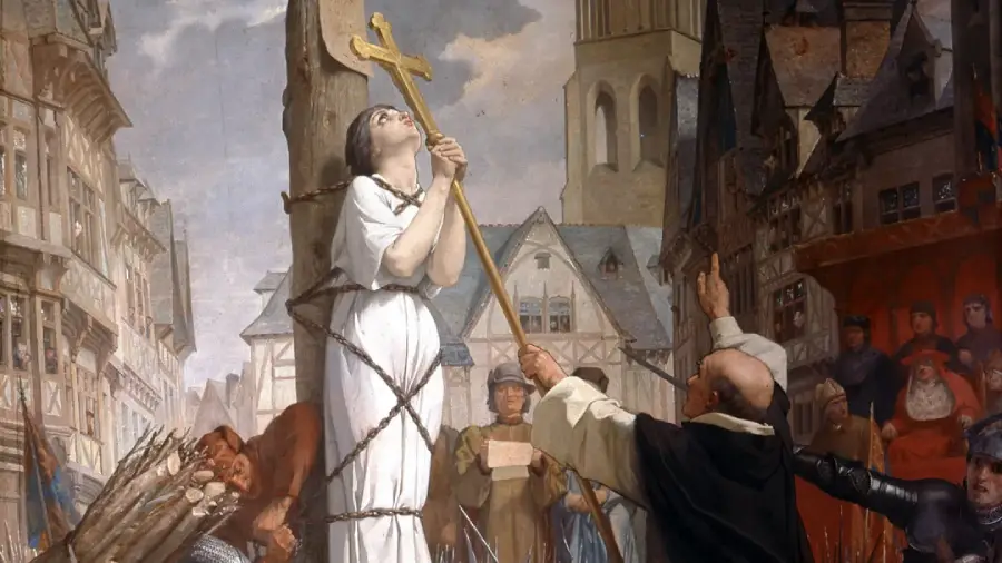 A young woman in white, tied to a wooden stake and holding a cross, looks upward as a crowd watches. Buildings and timber frame houses are in the background, and a man gestures toward her.