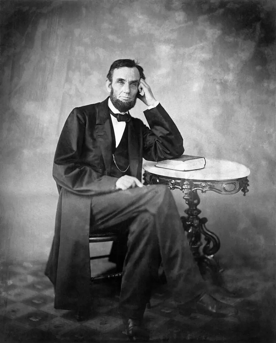 A bearded man in formal 19th-century clothing sits with his elbow resting on a small ornate table, hand to his face, beside a closed book on the table. The photo is black and white with a plain backdrop.