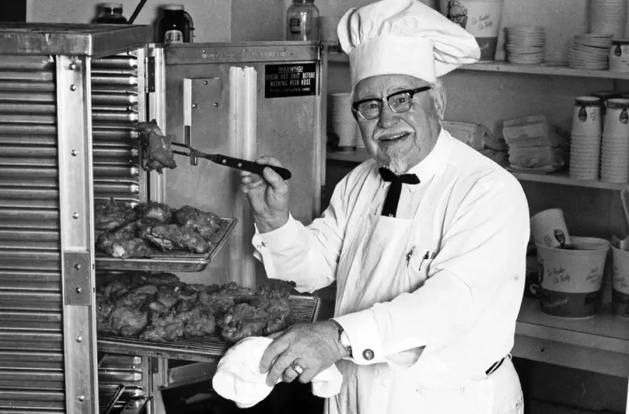 An elderly man in a chef's hat and glasses smiles while taking fried chicken from an oven rack in a kitchen, holding tongs in one hand and a towel in the other.