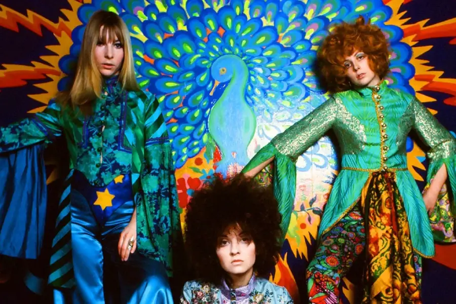 Three women in colorful, psychedelic 1960s fashion pose in front of a vibrant mural featuring a stylized peacock with radiating patterns in blue, green, orange, and yellow.