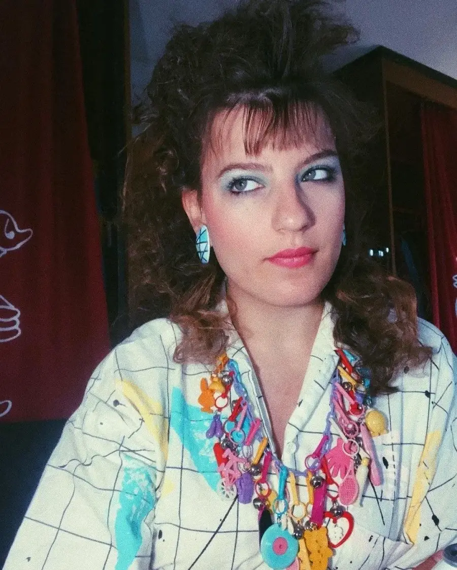 A woman with voluminous curly hair, bright makeup, geometric earrings, and a colorful necklace poses in a patterned shirt. The background features red curtains with white cartoon designs.