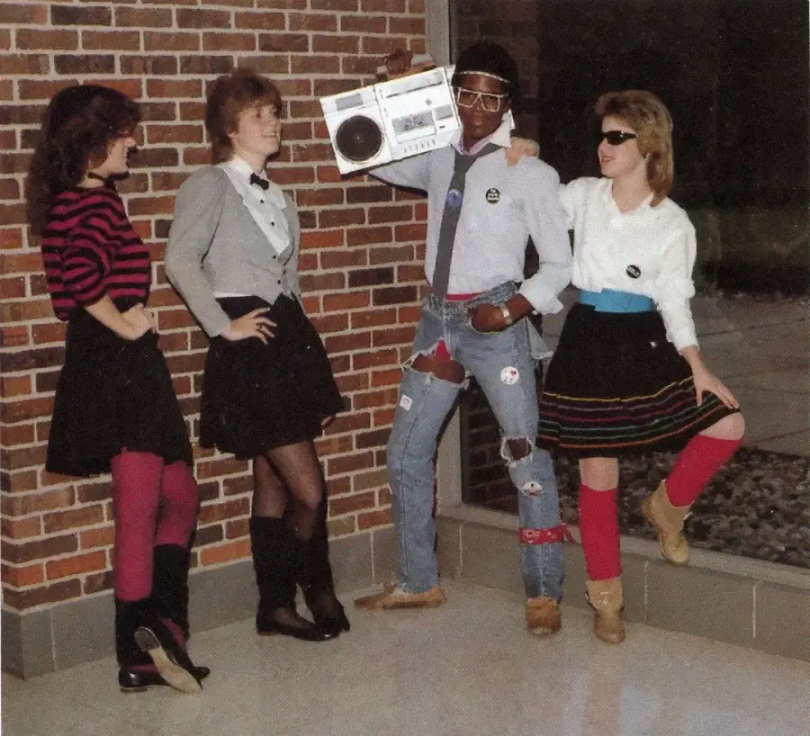 Four people dressed in colorful 1980s fashion stand by a brick wall. One holds a boombox on his shoulder, while the others pose confidently, wearing skirts, leggings, sunglasses, and boots.