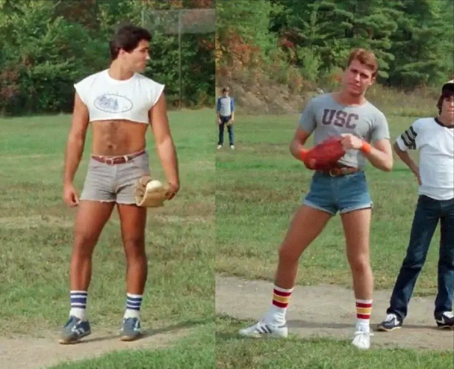 Two men stand on a grassy field wearing short shorts, tall striped socks, and athletic tops. One holds a baseball glove and the other holds a football. Trees are in the background and a few people are standing behind them.