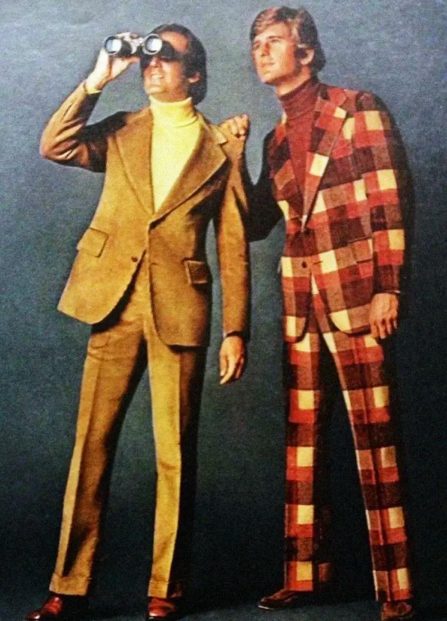 Two men in 1970s-style suits stand side by side; one wears a solid tan suit and yellow turtleneck while using binoculars, the other wears a bold red and brown plaid suit with a maroon turtleneck.