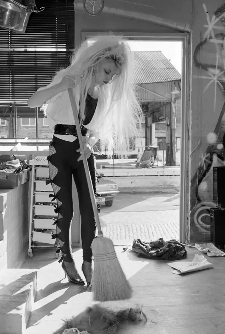 A person with long, voluminous hair wearing high heels and unique cut-out pants sweeps the floor inside a building, with sunlight streaming in through an open door.