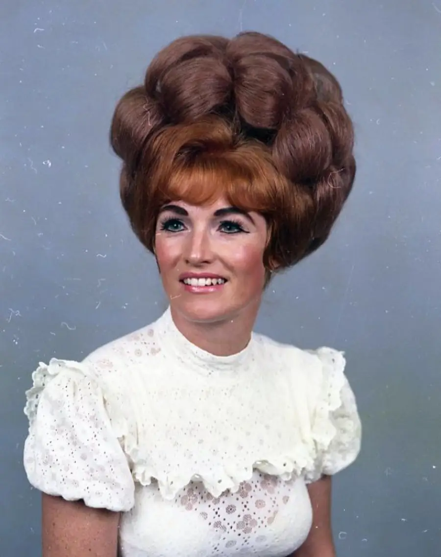 A woman with a large, voluminous beehive hairstyle and light red hair wears a white, lacy, short-sleeved blouse. She is smiling and posed against a plain light blue background.