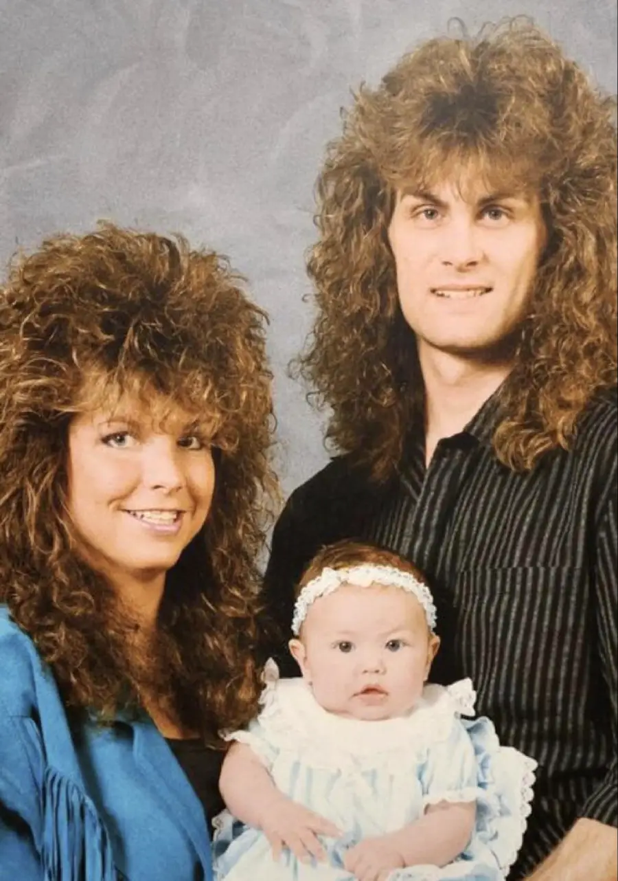 A woman and man with big, voluminous 1980s-style hair pose together, smiling, while holding a baby dressed in a light blue dress and white headband in a studio portrait with a gray backdrop.