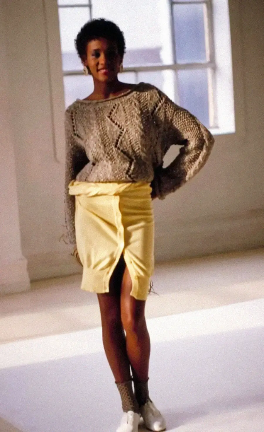 A woman stands indoors wearing a textured gray sweater, a high-waisted yellow skirt with a front slit, gray socks, and white shoes. She is posing with one hand on her hip in a sunlit room with large windows.