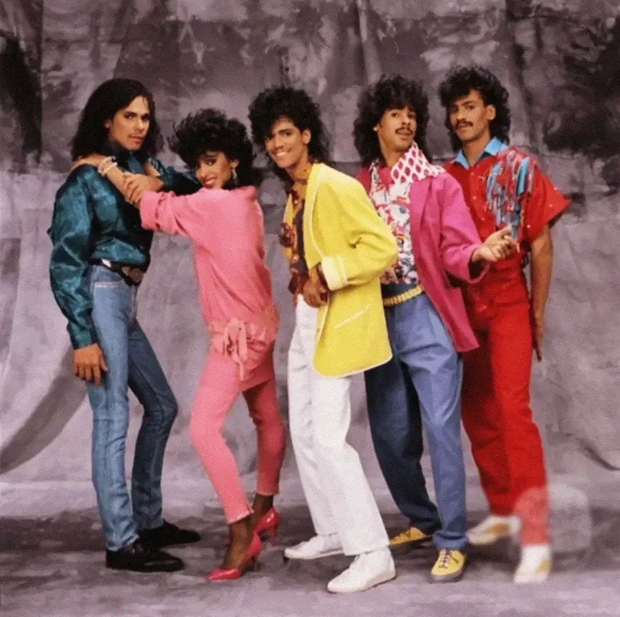 Five people pose in colorful, retro 1980s outfits against a gray studio backdrop. They smile and stand in a playful, dynamic formation, showcasing bright pink, yellow, blue, and red clothing with big, styled hair.