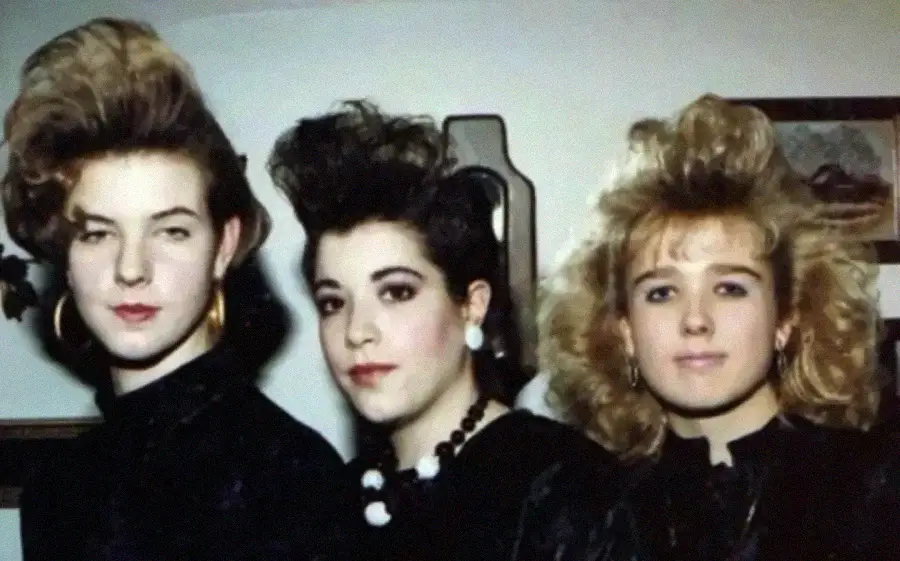 Three young women with voluminous, teased 1980s hairstyles and bold accessories, including large earrings and a beaded necklace, pose indoors against a plain background.
