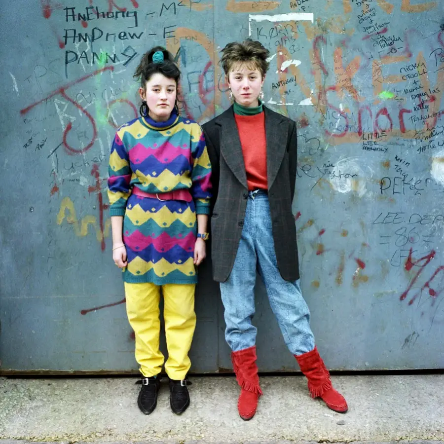 Two young people in colorful, bold 1980s-style outfits stand against a graffiti-covered wall. One wears a patterned sweater, yellow pants, and black shoes; the other sports jeans, a red shirt, blazer, and red boots.