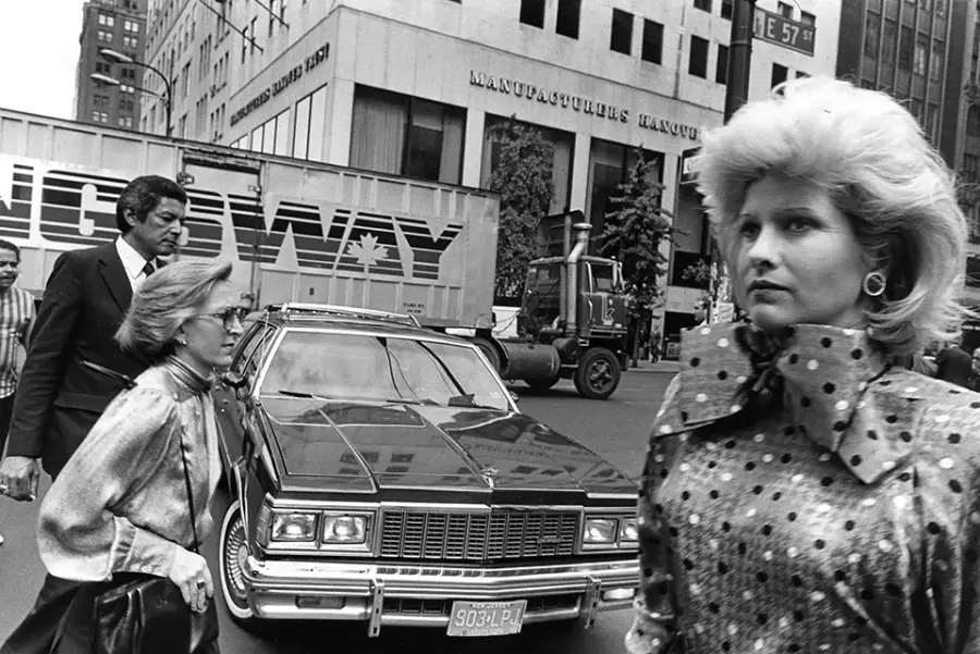 Black and white photo of people crossing a busy city street in front of a car; buildings, a truck, and a “Broadway” sign are visible in the background. One woman in polka dots is in the foreground.
