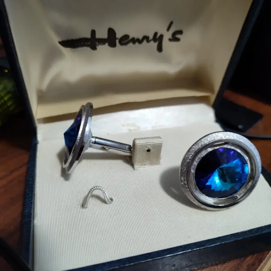A pair of silver cufflinks with large blue gemstones displayed in an open cream-colored box labeled "Henry's." One cufflink is upright, the other lies flat, with a small loose piece in front of them.