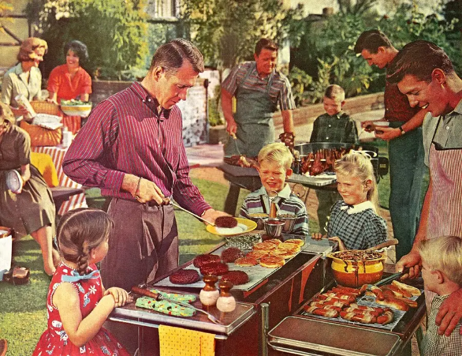 A group of adults and children gather outdoors for a backyard barbecue. A man serves burgers from a grill as kids line up with plates. Others chat and prepare food at picnic tables on a sunny day.
