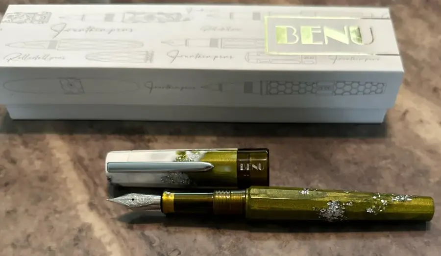 A green and silver Benu fountain pen with a decorative pattern rests on a marble surface, with its box featuring pen illustrations and the "BENU" logo in the background.