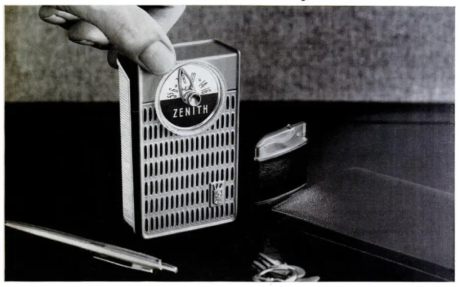 A hand holds a vintage Zenith portable radio above a table with a pen, keys, and a small case nearby. The radio has a silver grill and a round analog dial.