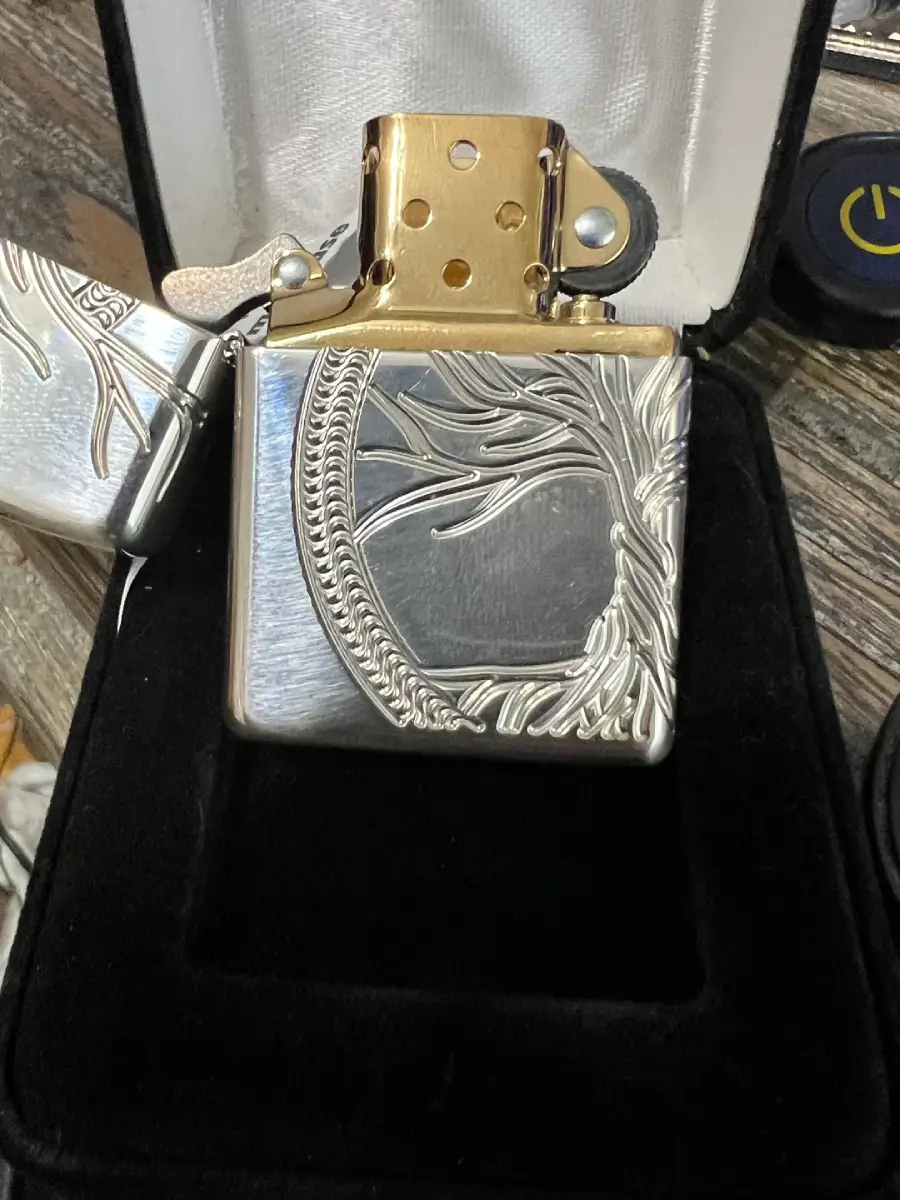 A silver and gold lighter with intricate braided and branch-like engravings is displayed upright in a black velvet box. The lighter’s lid is open, revealing its golden interior.