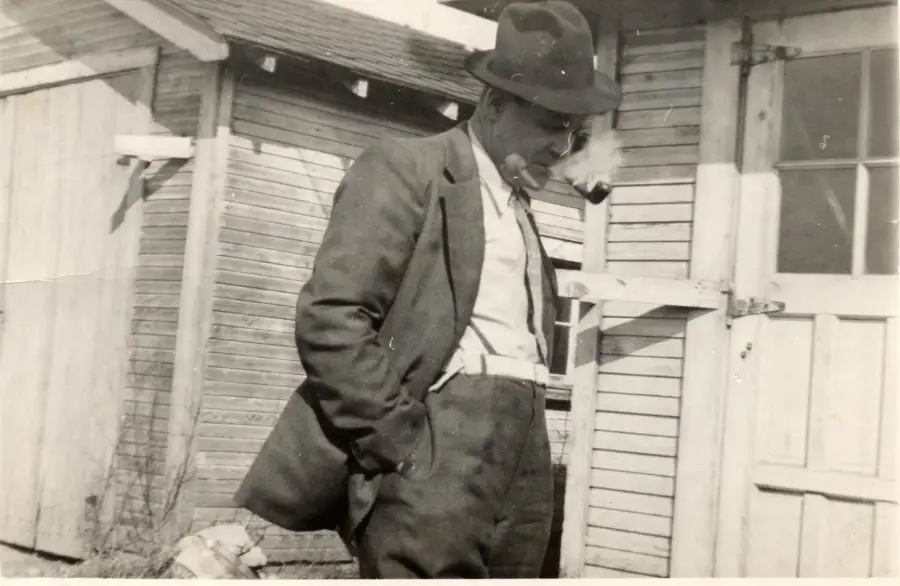 A man wearing a suit, tie, and fedora hat stands outside by a wooden building, leaning slightly forward with hands in his pockets and smoking a pipe. The photo is black and white, giving it a vintage feel.