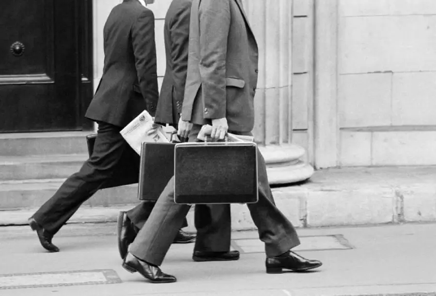 Three people in suits walking on a sidewalk, each carrying a briefcase. One person is also holding a folded newspaper. The image is black and white and shows them from the waist down.
