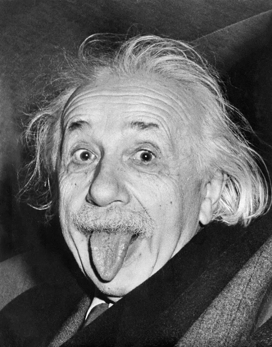 An older man with wild hair and a mustache playfully sticks out his tongue while looking at the camera in a black and white photo.