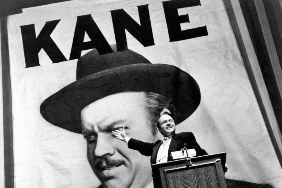 A man stands at a podium, gesturing toward a giant poster behind him that shows his face wearing a hat and the word "KANE" in bold letters above.