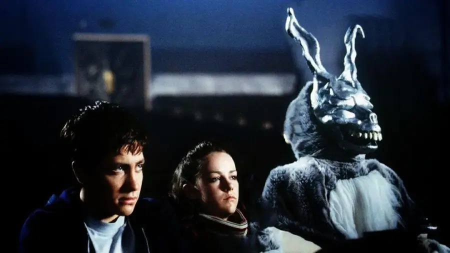 A young man and a young woman sit side by side in a dark theater, with a large, eerie figure in a creepy rabbit costume seated next to them. The mood is tense and mysterious.