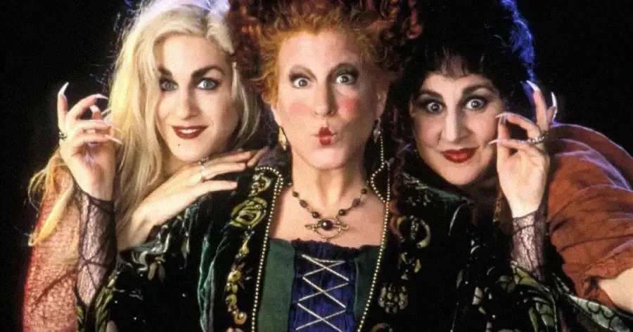 Three women in colorful, elaborate witch costumes pose together, with dramatic makeup and expressive faces. The woman in the center has red hair styled high, flanked by blond and dark-haired women, all with playful expressions.