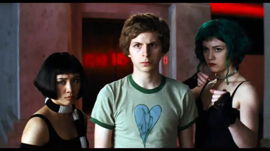 Three people stand together indoors. The person in the middle has short brown hair and wears a green t-shirt with a blue heart. The people on either side have short, dark hair and wear black outfits with intense expressions.