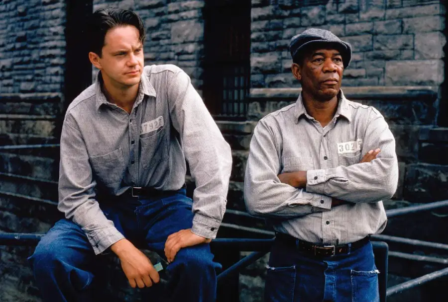 Two men in prison uniforms stand outside against a stone wall. One is sitting and the other is standing with arms crossed. Both look serious and are wearing gray shirts with number tags and blue jeans.