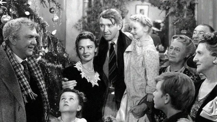 A group of people, including men, women, and children, gather around a decorated Christmas tree, smiling and looking happy in a cozy, festive setting; the scene is in black and white.