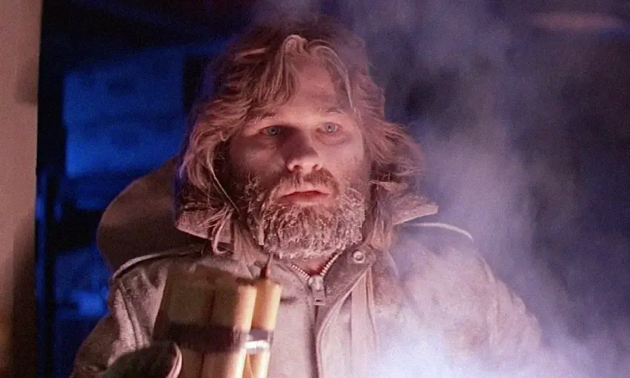 A bearded man in a heavy winter coat holds a bundle of dynamite sticks, surrounded by smoke and blue-tinted light, looking intensely ahead.