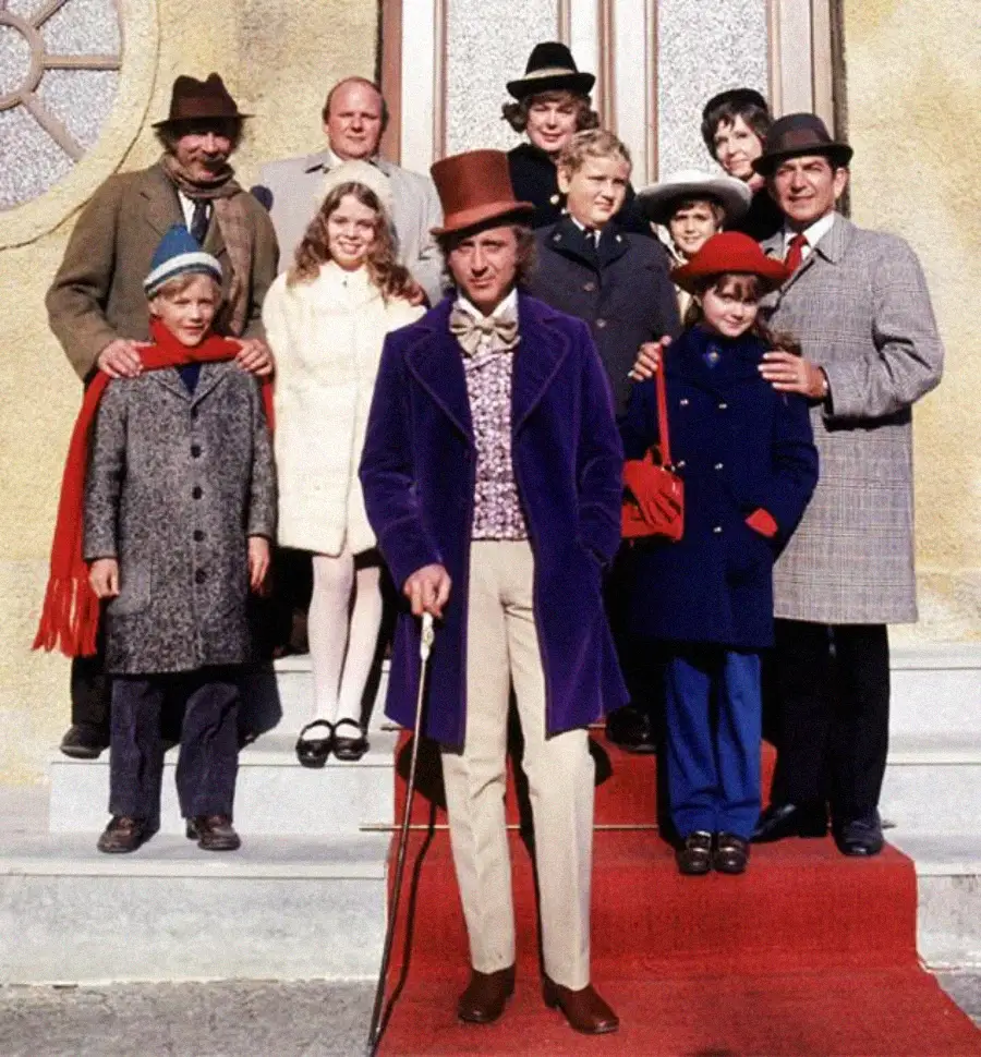 A group of people, including a man in a purple coat and top hat with a cane, stand on steps in front of a building. Several children and adults in colorful, vintage clothing pose together, smiling.
