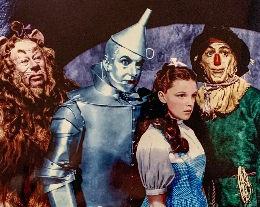 Dorothy, the Cowardly Lion, the Tin Man, and the Scarecrow stand together in colorful costumes from the movie "The Wizard of Oz," posing against a dark background.