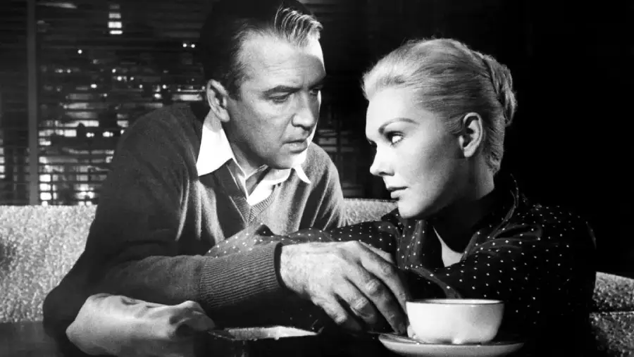 A man and a woman sit closely together at a table in a dimly lit room. The man looks intently at the woman, who gazes thoughtfully downward, her hand resting near a cup of coffee in front of her.