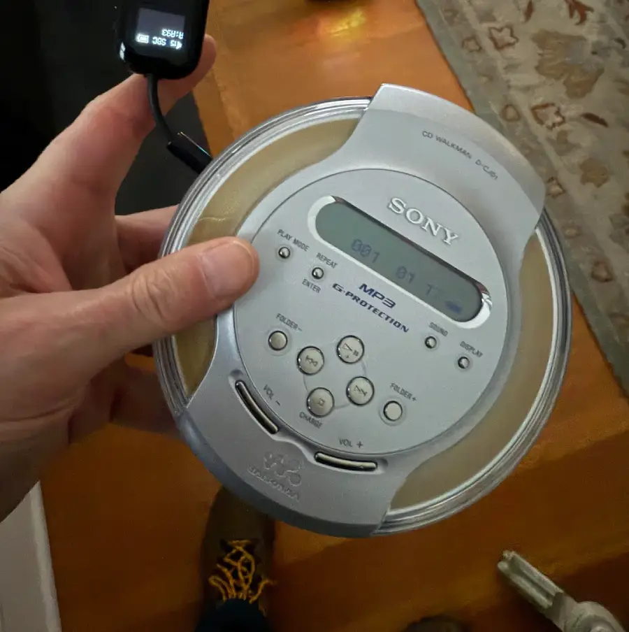 A hand holds a silver Sony CD Walkman portable music player with visible buttons and display. Part of a carpet and wooden floor are seen in the background.