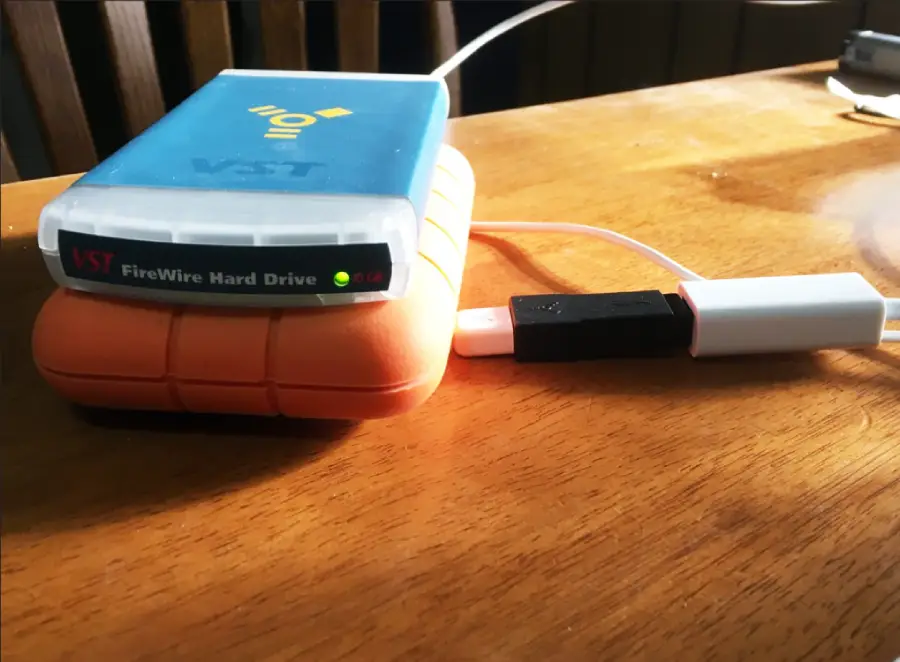 Two external hard drives, one blue labeled "VST FireWire Hard Drive" and one with an orange bumper, are connected by cables on a wooden table in sunlight.