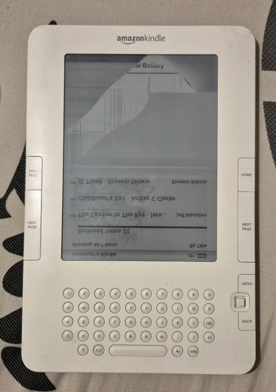 A white Amazon Kindle e-reader with a cracked and malfunctioning screen; part of the display is distorted with lines, and the text appears upside down and unreadable. The physical keyboard and navigation buttons are visible.
