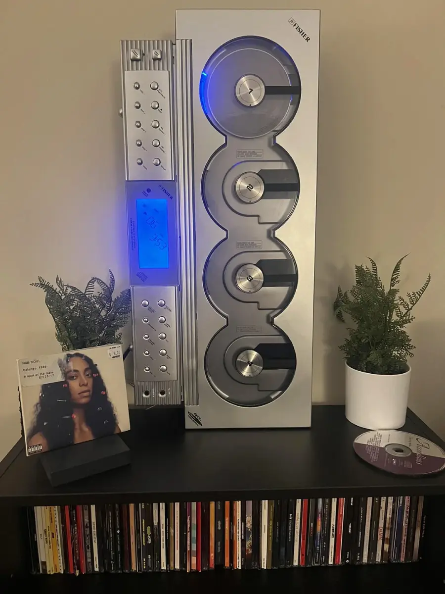 A modern vertical CD player with a blue light is on a black shelf, flanked by two potted plants. Below the player are CDs and a Sade album on display, with an open CD case and disc to the right.