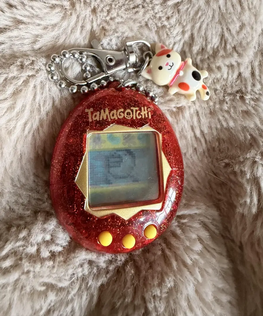 A red glittery Tamagotchi keychain with yellow buttons and a small cat charm rests on a soft, beige furry surface. The Tamagotchi screen displays pixel graphics.