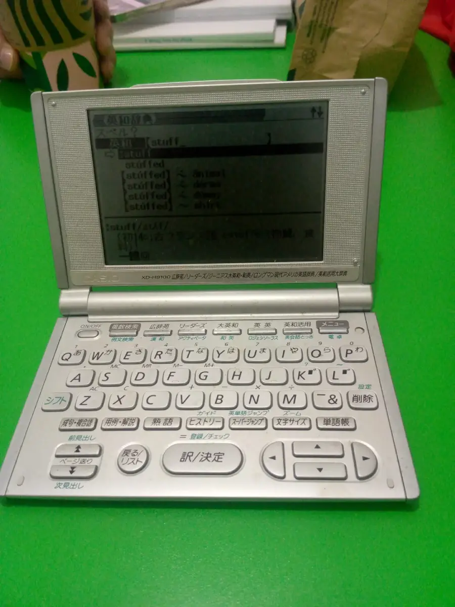 A Japanese electronic dictionary with a QWERTY keyboard sits open on a bright green table. A hand holding a drink cup and a brown paper bag are visible in the background.