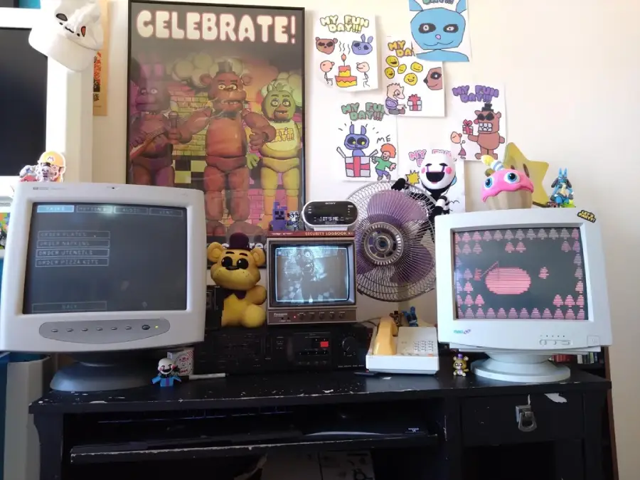 A desk with two old computer monitors, a vintage TV, and various Five Nights at Freddy's toys and art. The wall behind displays colorful fan art and a large "Celebrate!" FNaF poster. The setup is playful and nostalgic.