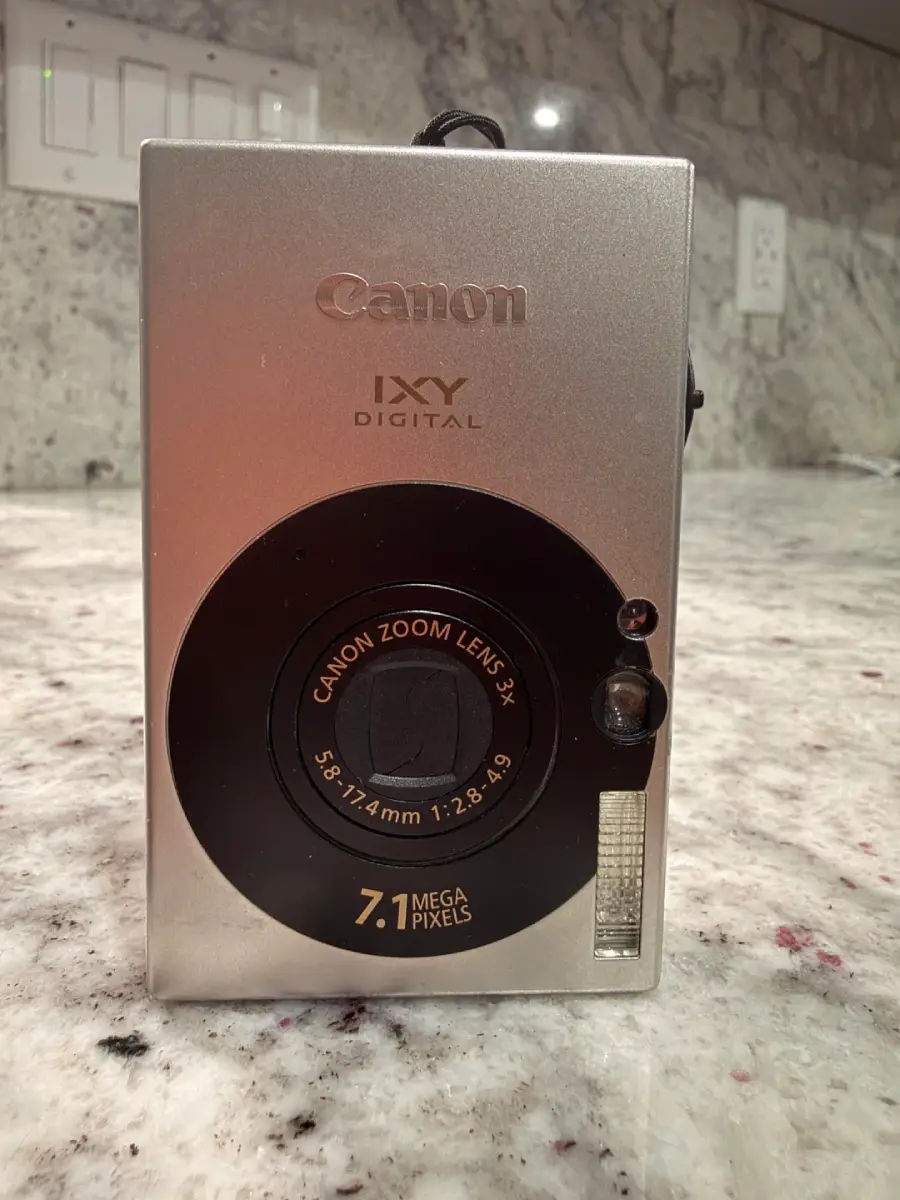 A silver Canon IXY Digital compact camera with a black circular lens and "7.1 Mega Pixels" written on the front, placed on a speckled granite countertop with a tiled backsplash in the background.