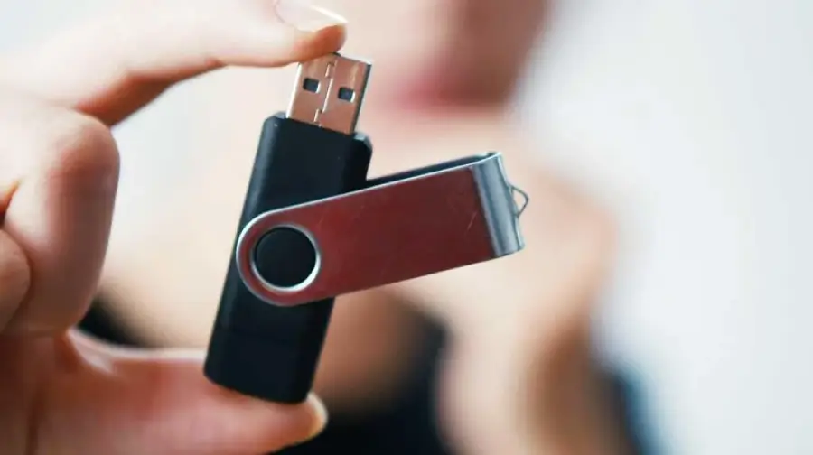 A hand holding a black USB flash drive with a metal swivel cover, shown against a blurred background.