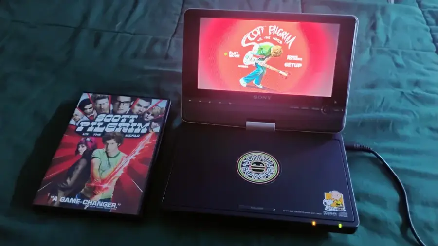 A portable DVD player displays the Scott Pilgrim vs. the World DVD menu. Next to it is the movie’s DVD case on a blue-green bedspread. The device has stickers on it and is plugged in.
