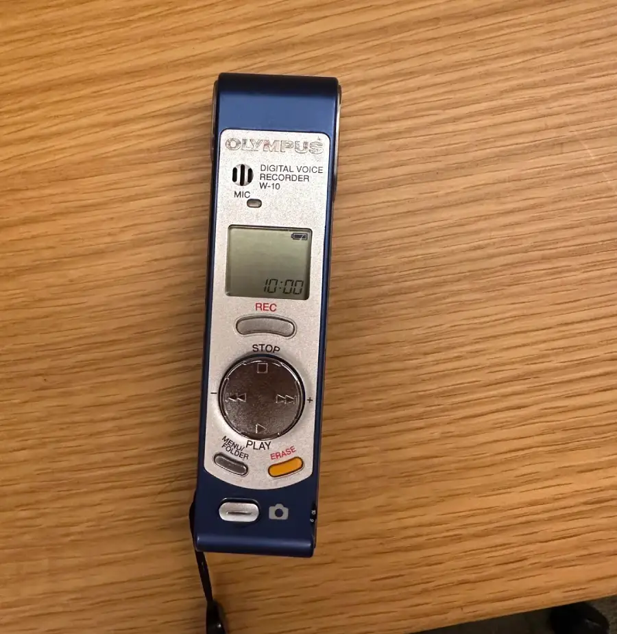 A silver and blue Olympus digital voice recorder is placed on a wooden surface, with REC, STOP, and PLAY buttons visible along with a small digital display screen.